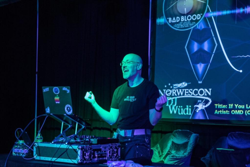 Me lit in blues and greens, behind my laptop and DJ equipment and in front of a projection screen with graphics and my DJ Wüdi at Norwescon logo, gesturing and singing along with the song being played.