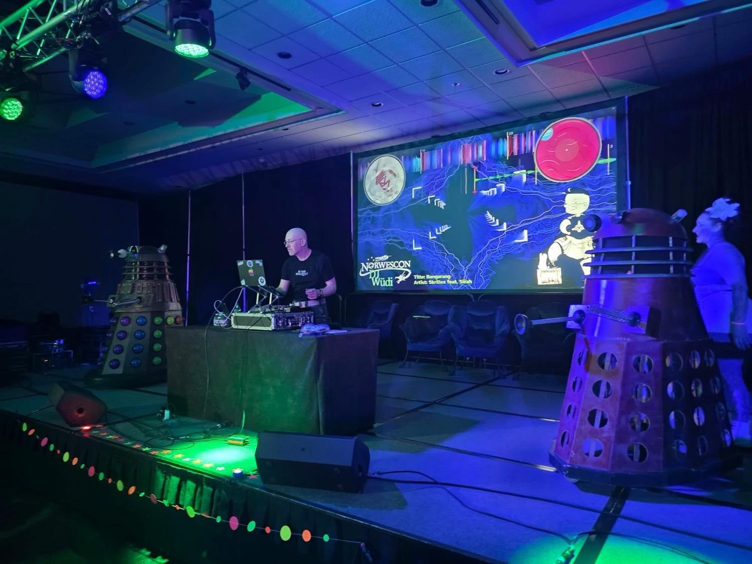 Me on stage behind table with my DJ equipment and in front of a large screen with fancy graphics, flanked by two Daleks.