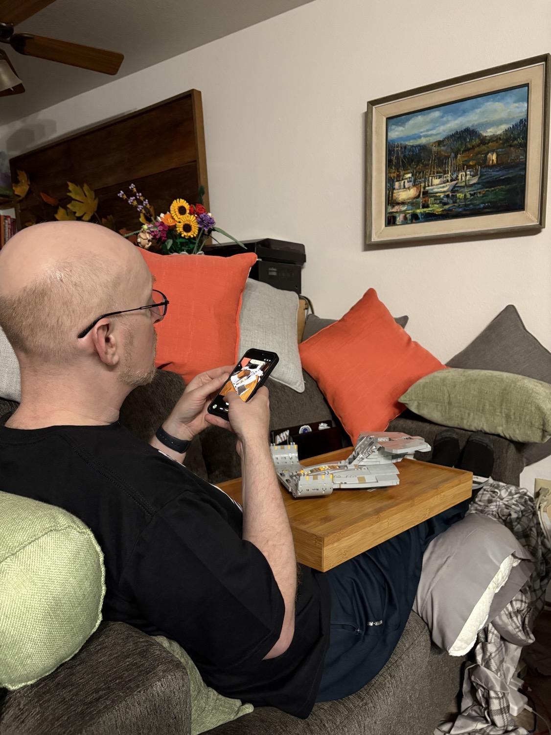Me sitting on our couch with a wooden TV tray on my lap. On the TV tray is a partially completed Lego starship Enterprise; I'm in the midst of taking a photo of it with my iPhone.