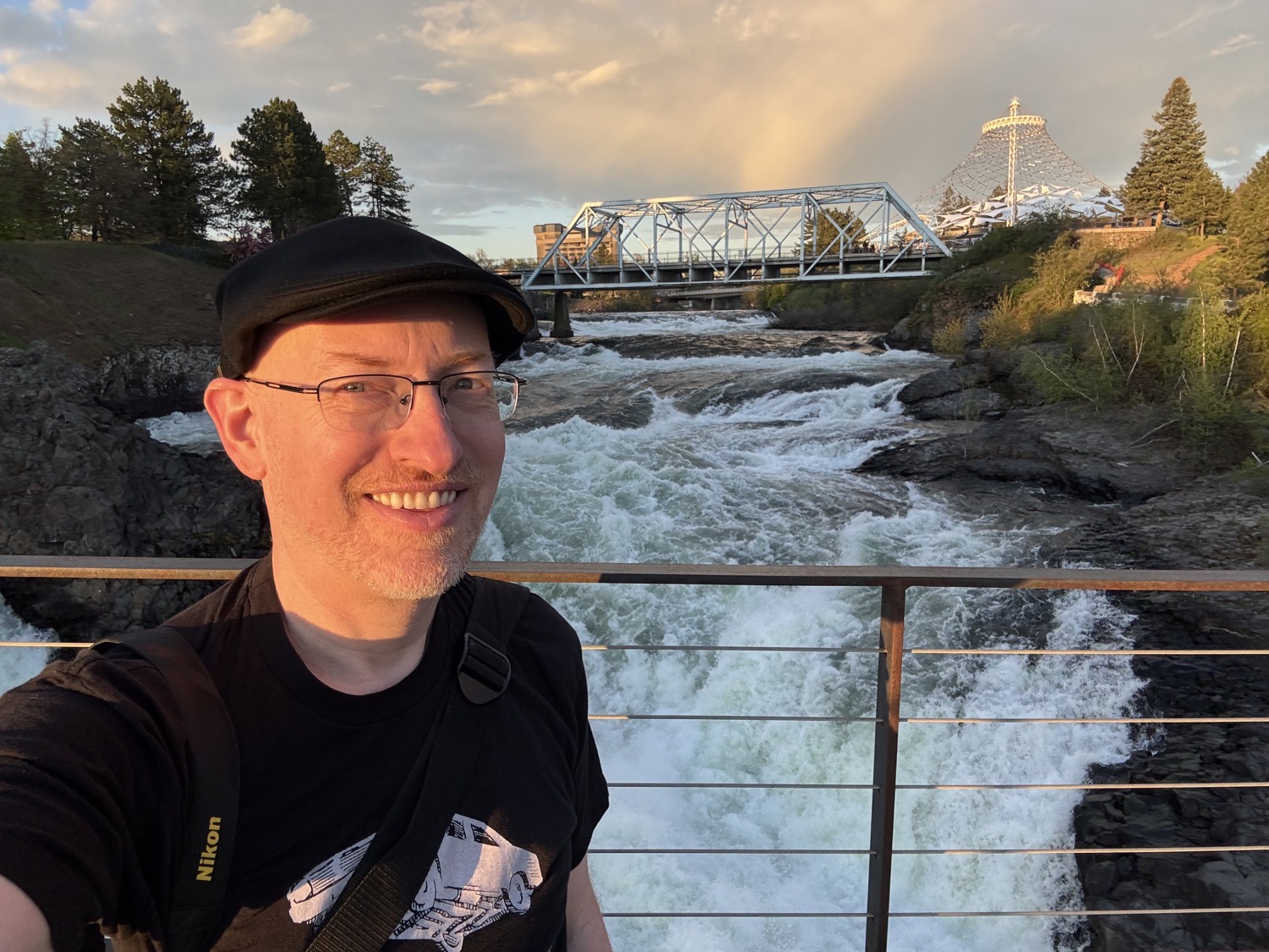 Me lit by the setting sun, standing on a bridge over a rushing waterfall, with another bridge and a Spokane park in the background.