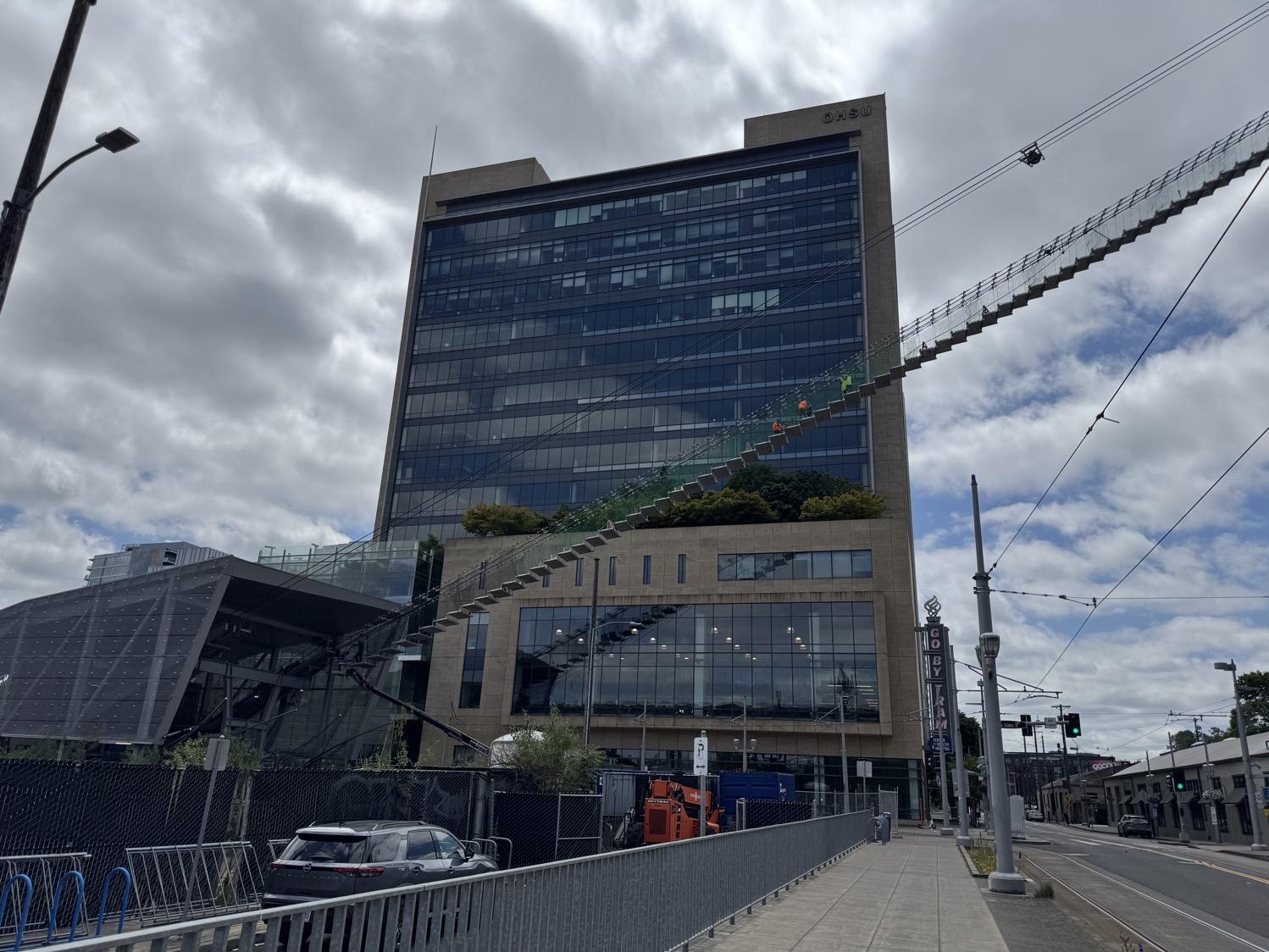 In front of an office building, wires for a tram line go diagonally upwards across the shot. One set of wires has temporary netting and stairway platforms dangling from it as workers move along the suspended stairs.