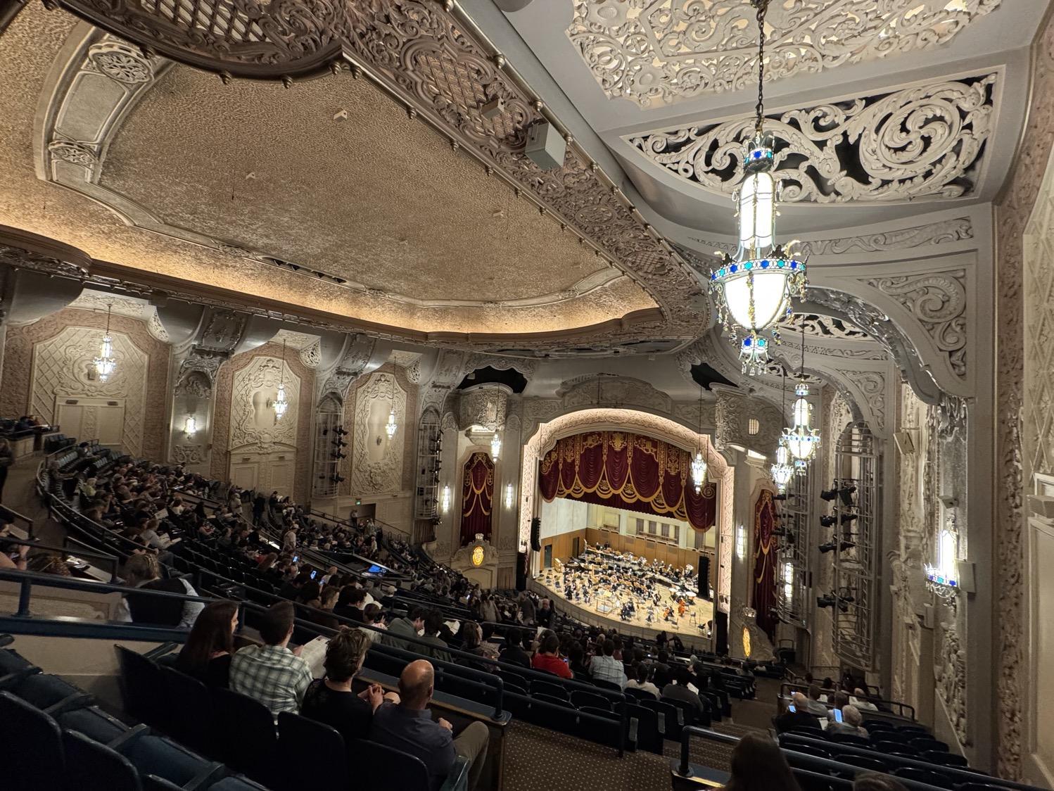 Wide angle view of an ornately decorated theater seen from the very rear of the uppermost balcony. The walls are white and beige, ornately carved, chandeliers with blue and white gland hang from the ceiling, and red and gold curtains frame the stage in the distance, with a symphony starting to take their seats on the stage.