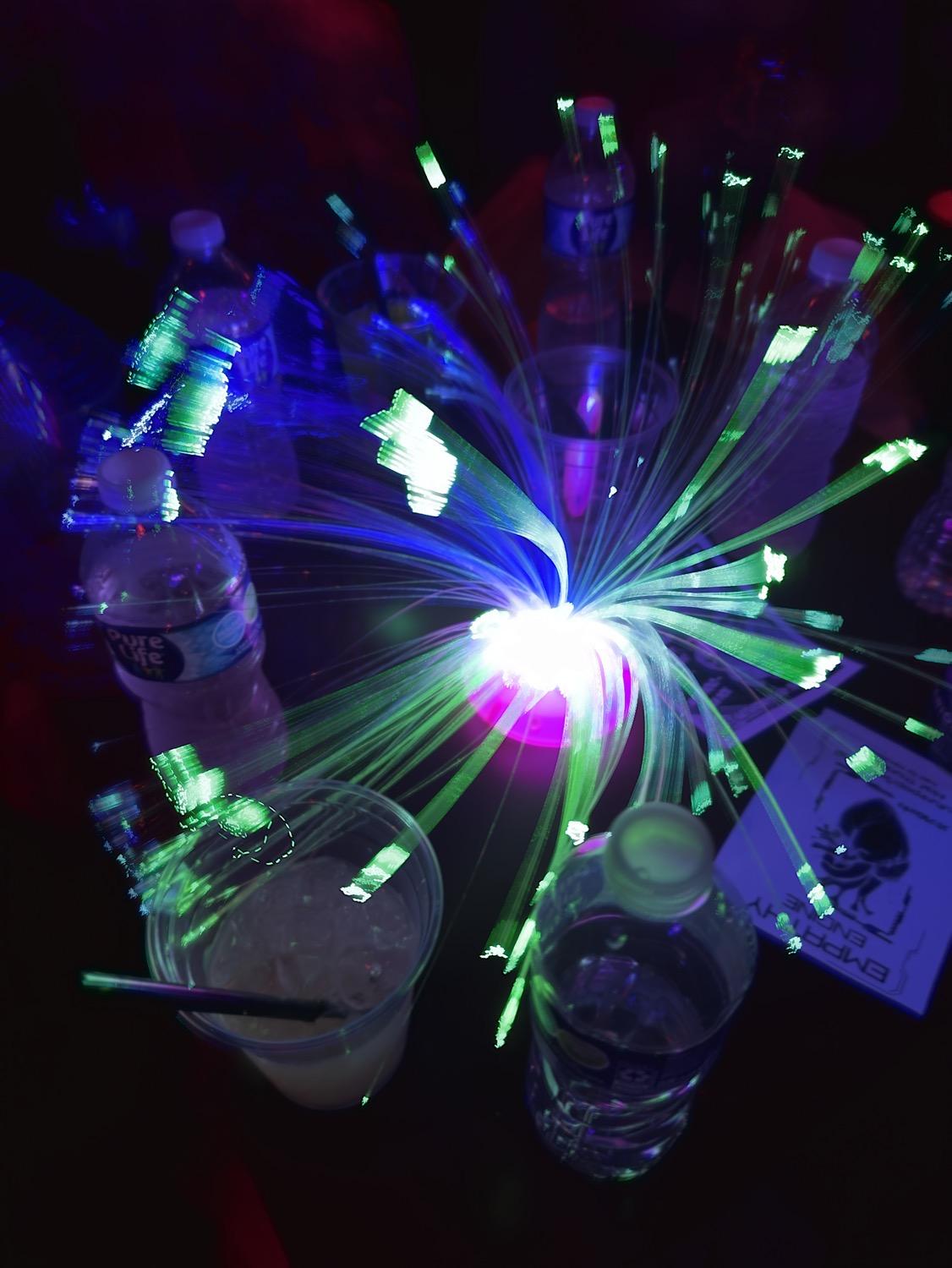 On a dark table surrounded by drinks and water bottles, a small LED centerpiece with fiber optic strands glowing in green, slightly blurred as the fiber optic strands move slightly.