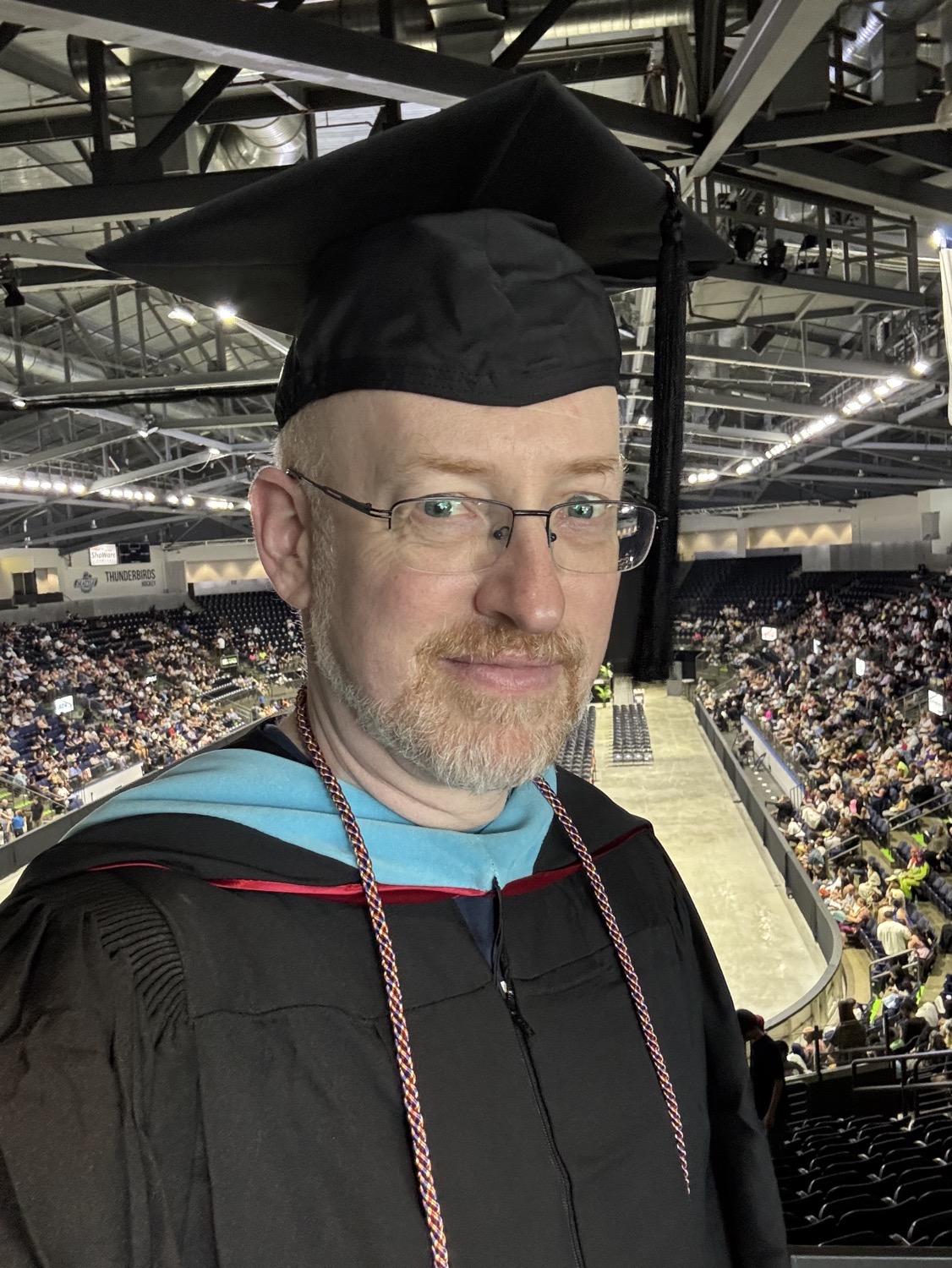 IMG_6239.jpeg Me wearing the traditional black robe, black mortarboard cap, master's hood with blue and red trim, and a rainbow honors cord, with a stadium full of people in the stands visible behind me.