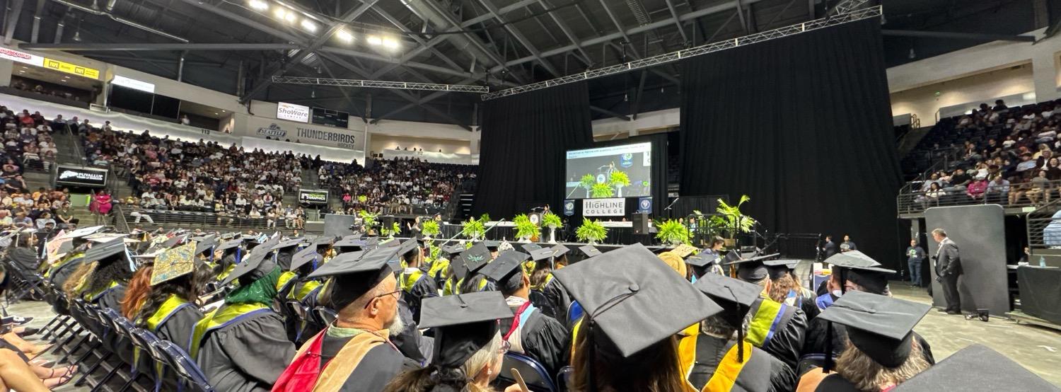 IMG_6246.jpeg Panoramic shot from the floor of the stadium mid-ceremony, with faculty and graduates in their robes seated in front of a stage with a large video screen showing the current speaker.