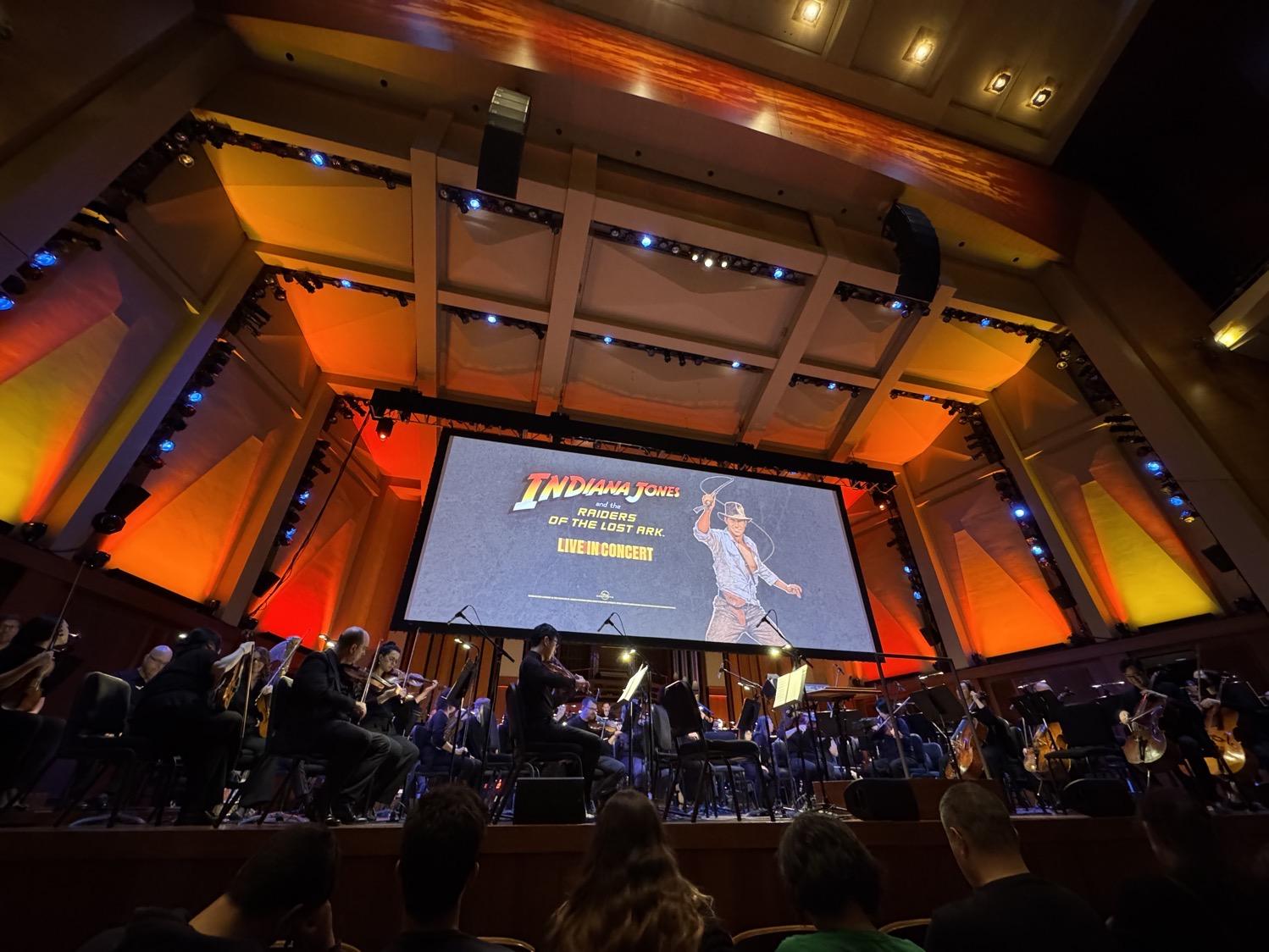 The stage at Seattle's Benaroya Hall, lit is reds, oranges, and golds, with the symphony under a large screen showing the title card for Raiders of the Lost Ark Live in Concert.