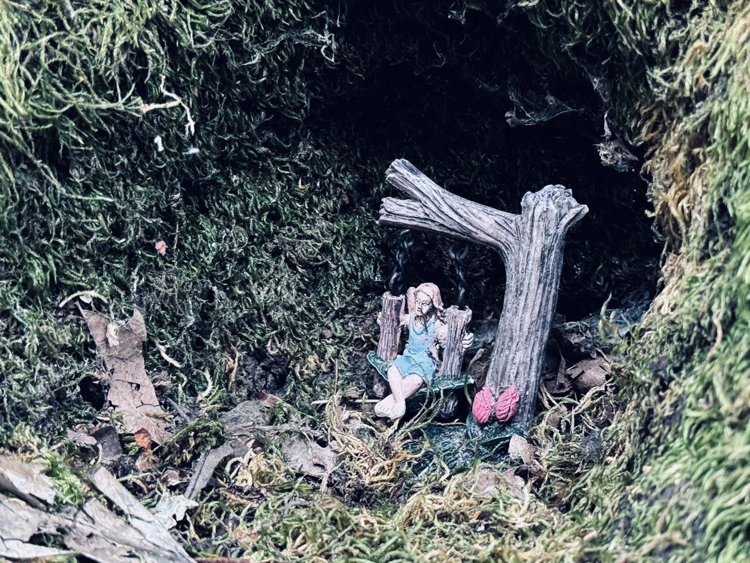 IMG_6320.jpeg A small sculpture of a fairy sitting on a swing made of twigs and leaves hanging from a tree branch, placed in a moss-covered hollow in a tree.