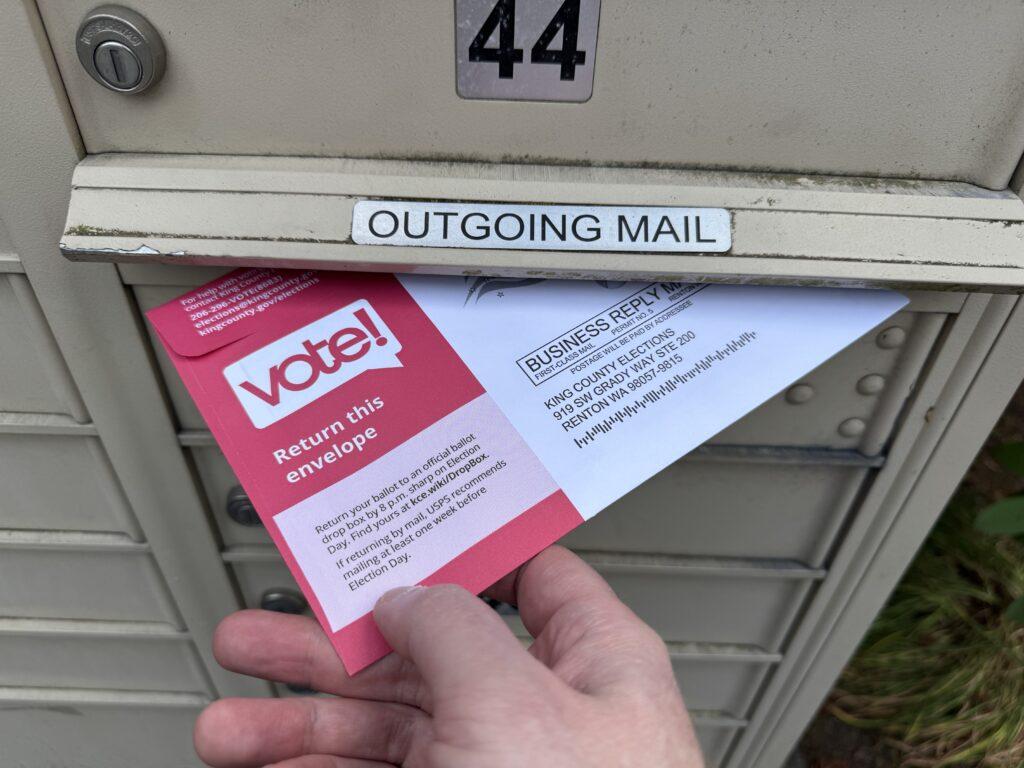 My hand placing a Washington state vote by mail return envelope into a mailbox.