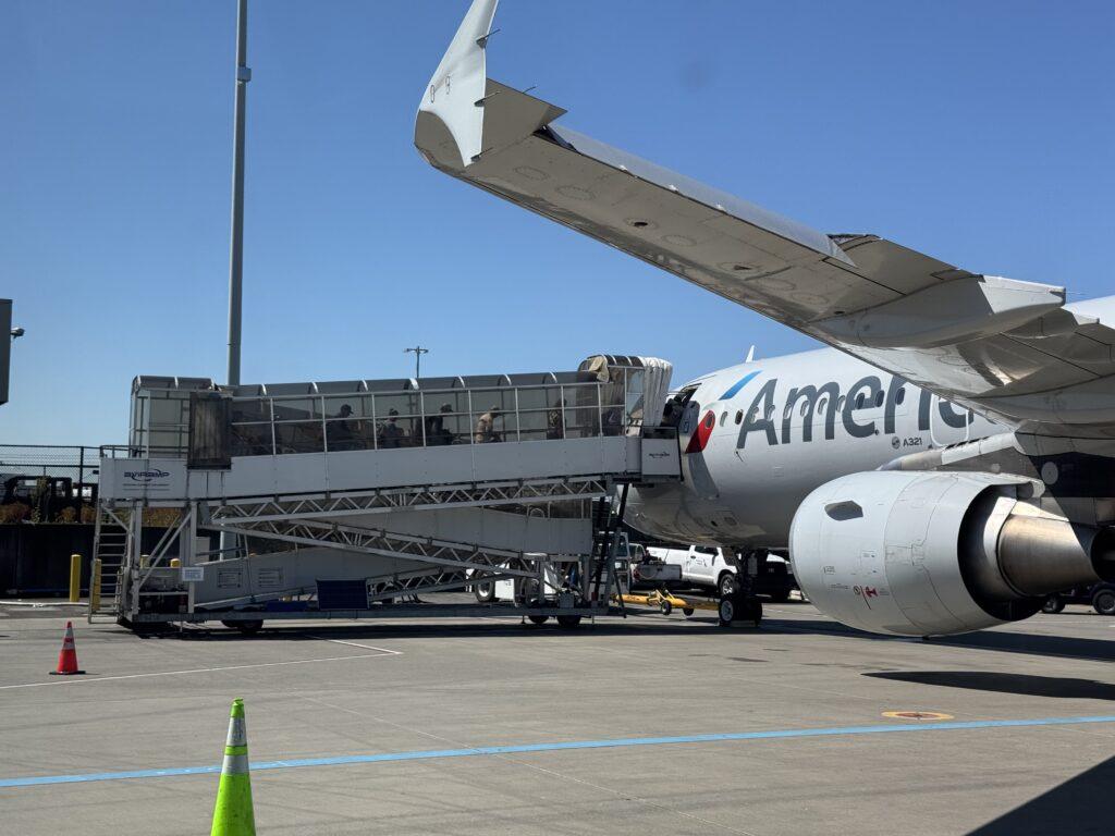 An American Airlines plane sitting on the tarmac with a mobile ramp platform letting passengers embark.