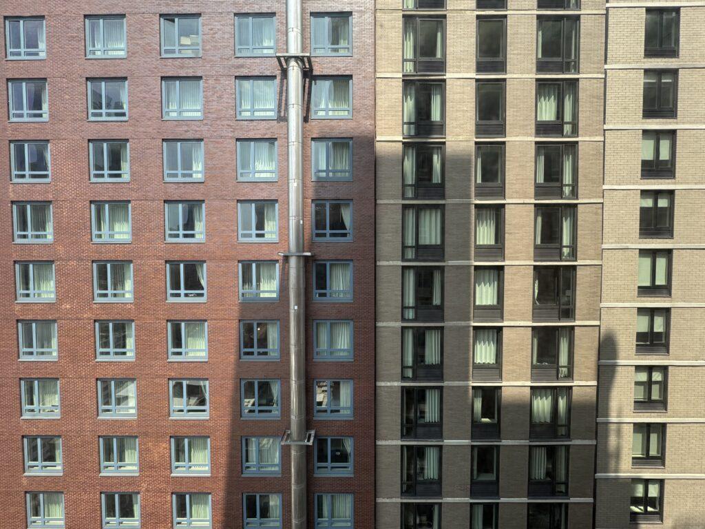 A grid of windows of two buildings side-by-side, one a darker brown and one a lighter tan, with a large metal tube running down the darker building.