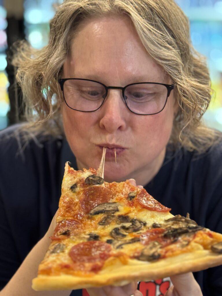 A white woman with short wavy blonde hair takes a bite from a large piece of pizza, with strings of cheese leading from the pizza to her pursed lips.
