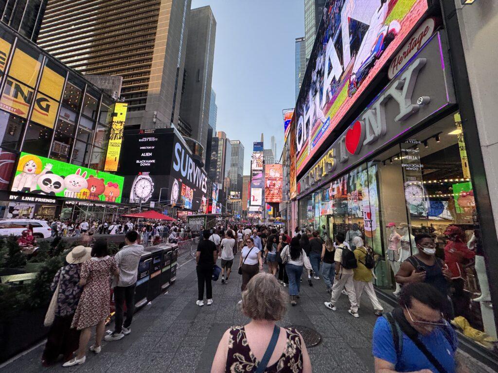 A wide-angle shot of a woman walking through downtown New York City into Times Square, surrounded by other people, giant flashing billboards, and tall buildings.