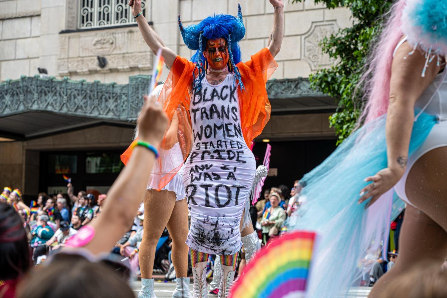 A drag queen with blue hair and red and black face paint, wearing an orange gauzy shawl and a white dress with black writing that says, 'Black trans women started Pride and it was a riot'.
