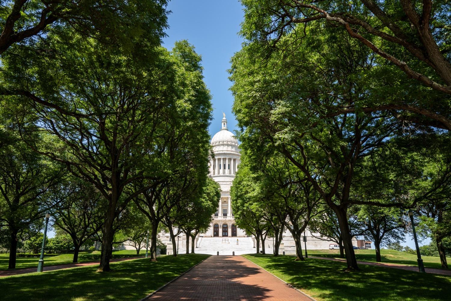 At AHEAD 2026- State House Tour - 115.jpg A red brick walkway shaded by tall green oak trees leads to the white marble staircase, edifice, and dome of a large governmental building under a clear light blue sky.