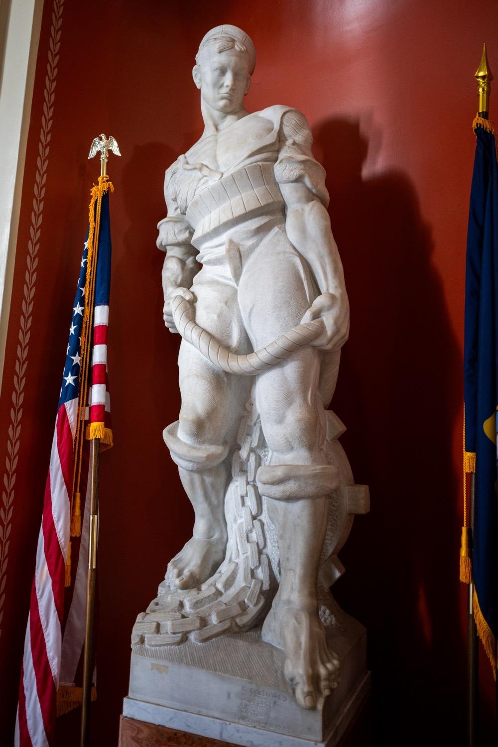 At AHEAD 2026- State House Tour - 143.jpg A white stone sculpture of a sailor, leaning back slightly on one leg, holding a rope in both hands, wearing a sweater that open and falling off of his shoulders and with the sleeves rolled up to expose a very muscular chest and arms, and with very tight pants rolled up at the cuffs to show off his muscular legs.