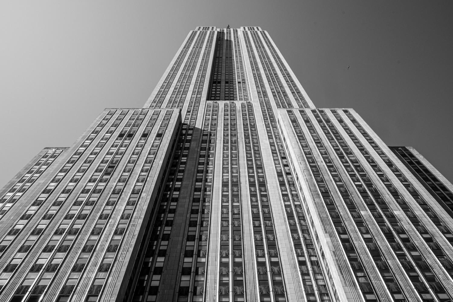 Exploring New York City - 369.jpg A black and white shot of the Empire State Building from street level, symmetrically framed, so that the straight lines of the building rise and converge towards the top center of the photo.