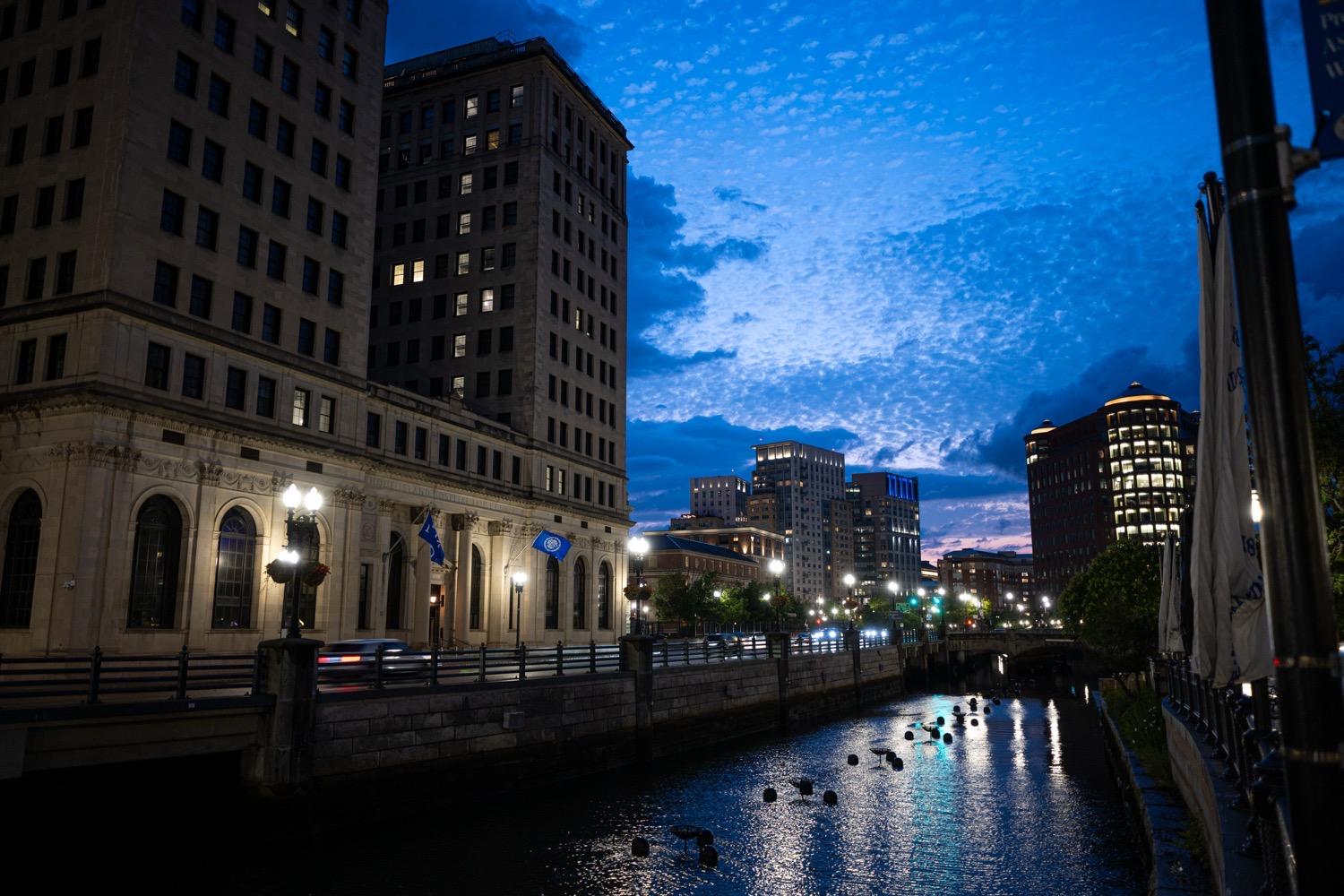 Exploring Providence - 110.jpg A night shot down the Providence river, which runs through downtown Providence. Tall stone buildings lit by streetlights flank a river banked by stone walls, under a deep blue evening sky that fades to a light purple at the horizon behind more city buildings. The sky and buildings are reflected in the surface of the river.