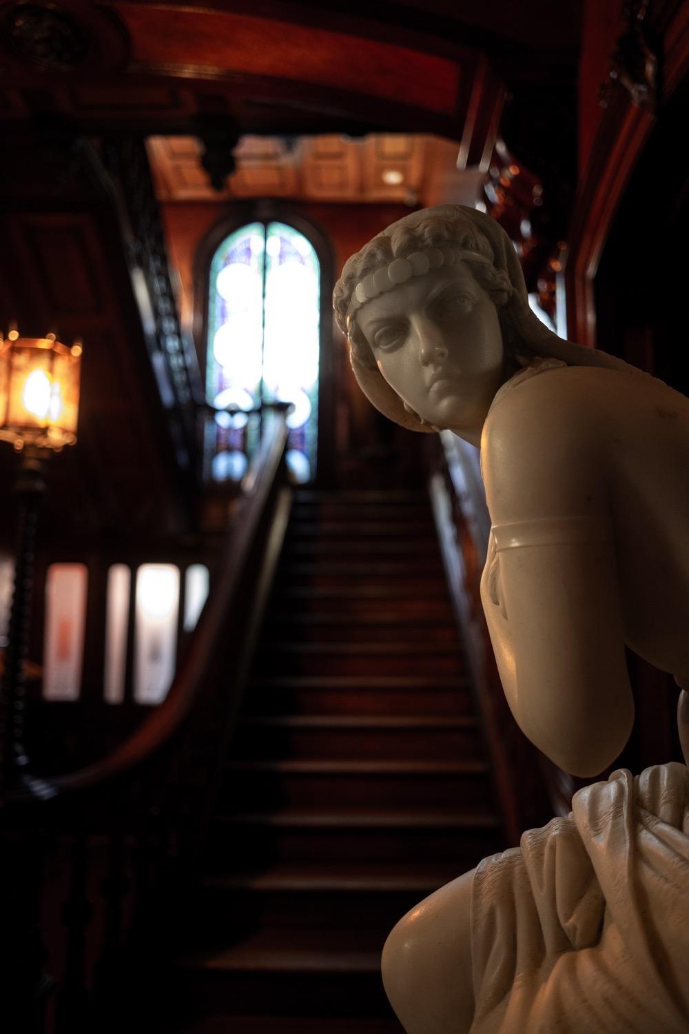 Exploring Providence- Lippett House Museum - 070.jpg With a slightly blurry dark wooden staircase in the background, a white stone sculpture of a crouching woman with a severe expression looks towards the camera as if angry at the photographer.