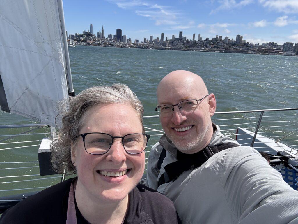 Selfie of my wife and I on a boat with a sail just visible to one side of us and the San Francisco skyline in the distance behind us.