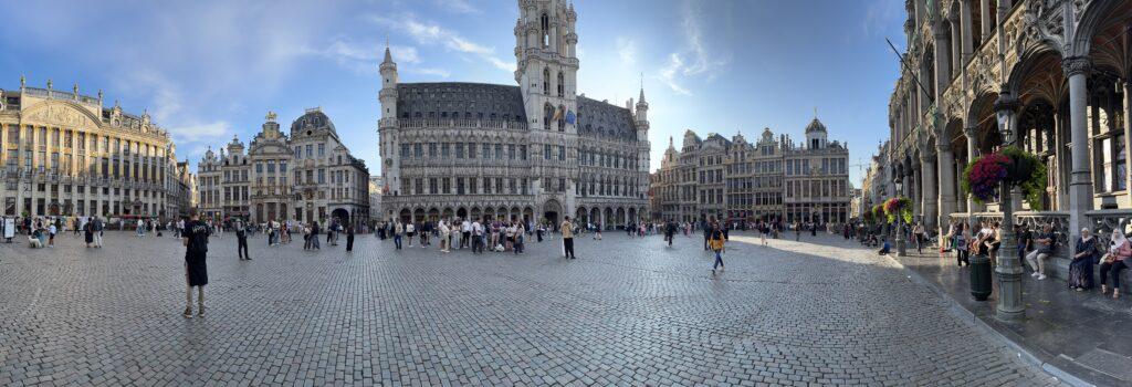 Panoramic view of a large cobblestoned square surrounded on all sides by large, ornate gothic and gold-trimmed buildings.