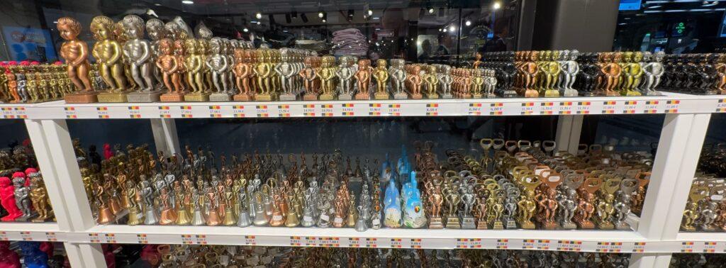 Two shelves of reproductions of the Mannekin Pis peeing boy statue, in various sizes and in gold, silver, or bronze colors, stacked several rows deep.
