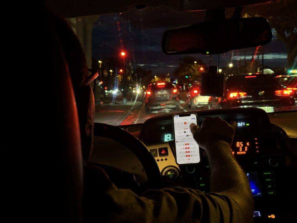 Shot from inside a cab, brake lights of nearby cars on a busy nighttime road can be seen through a windshield as the cabbie reaches over to scroll through sports scores on the screen of his dash-mounted cell phone.