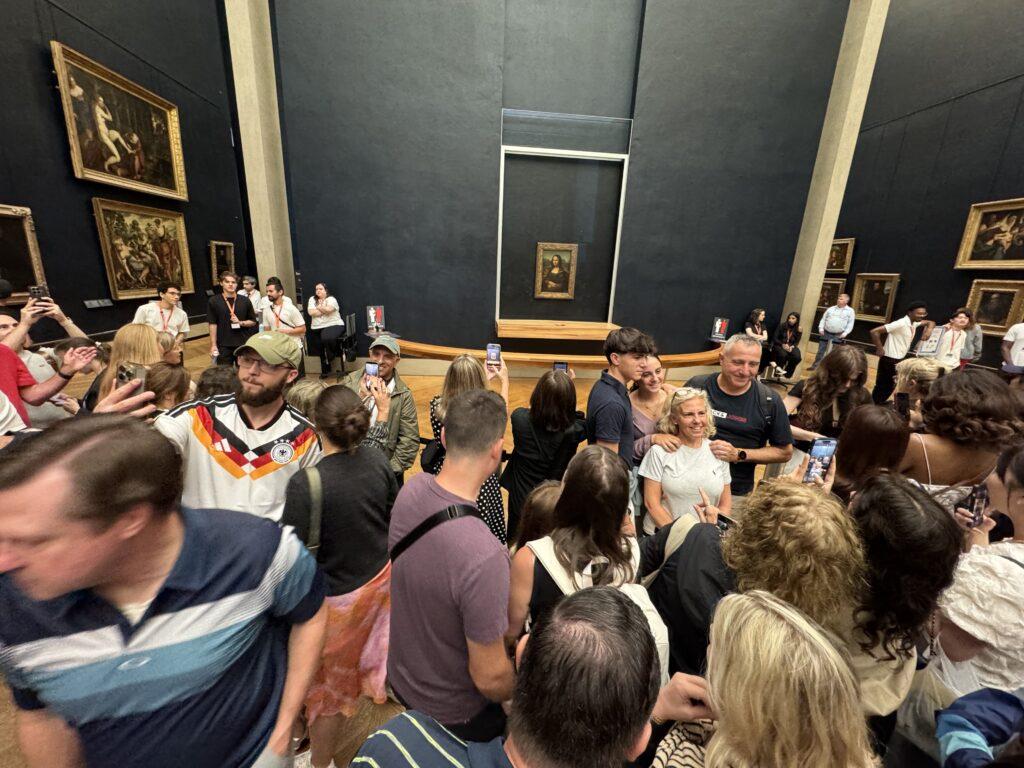 Wide-angle shot of a crowd of people standing behind a barrier, many with phones out to take photos, with the Mona Lisa alone in a dedicated alcove on a black wall.