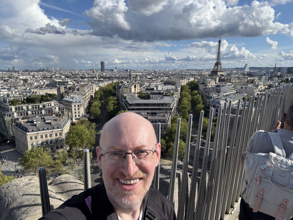 Selfie of me taken atop the Arc de Triomphe, with Paris behind me, the Eiffel Tower very visible towards the right side of the photo.