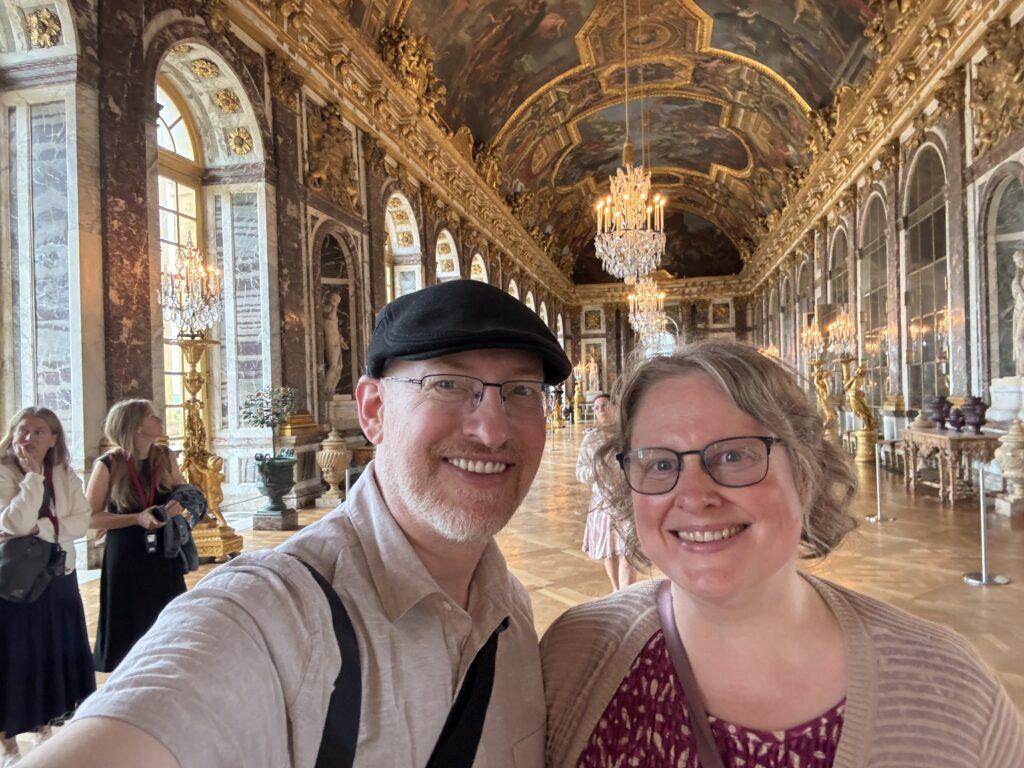 Selfie of my wife and I in the Versailles Hall of Mirrors, a long, ornately decorated and gilded room with painted ceilings, large windows on one side, and large mirrors on the other.