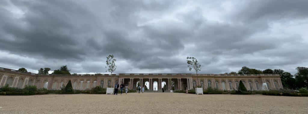 Panoramic shot of the Grand Trianon, a long, single-story marble building with a columned central walkway connecting two windowed wings, under a sky of dark grey clouds.