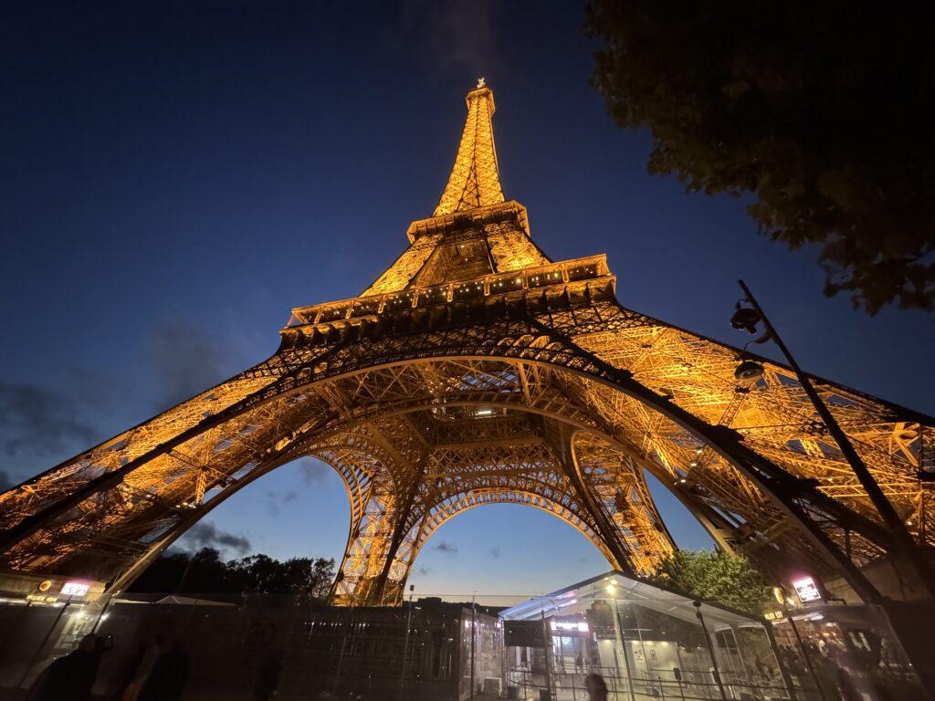 Wide-angle view of the Eiffel Tower as seen from the ground very close to the base, all lit in gold under a deep blue twilight sky, the lens distortion emphasizing the immensity of the structure overhead.