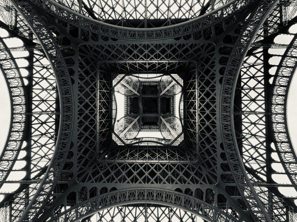 Black and white shot looking up the Eiffel Tower from dead center underneath it, forming a very geometric pattern of lines and angles made by the struts, beams, and girders of the tower’s metalwork.
