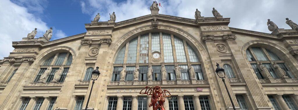 The front of the Gare du Nord train station, a stone and glass edifice with many statue figures between windows and along the roofline.