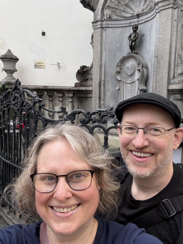 Selfie of my wife and I in front of a stone alcove with a fountain made of a small bronze sculpture of a small boy peeing.
