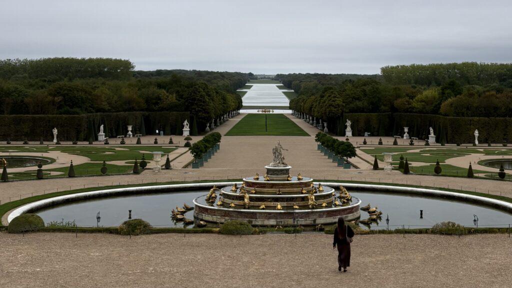 With an ornate fountain in the foreground and the water of the Grand Canal in the distance, the gardens of Versailles are seen early in the morning under a grey sky, with just one lone figure walking towards the fountain.