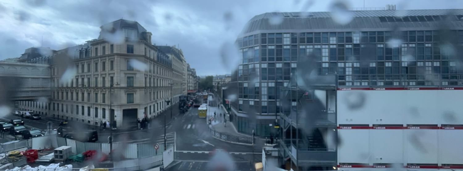 Panoramic view through a rain-dropped window of the Parisian street outside the train station, with an old style ornate stone building on one side of a street and a modern office building on the other.