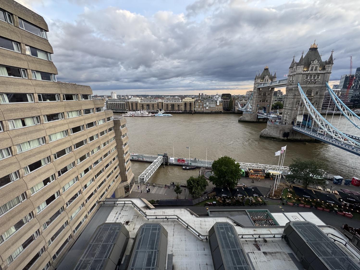 Wide-angle shot of the Thames river under a cloudy blue sky, with the Victorian stone-and-steel Tower Bridge to the right of the image and the Brutalist concrete-and-glass facade of the Tower Hotel on the left of the image.