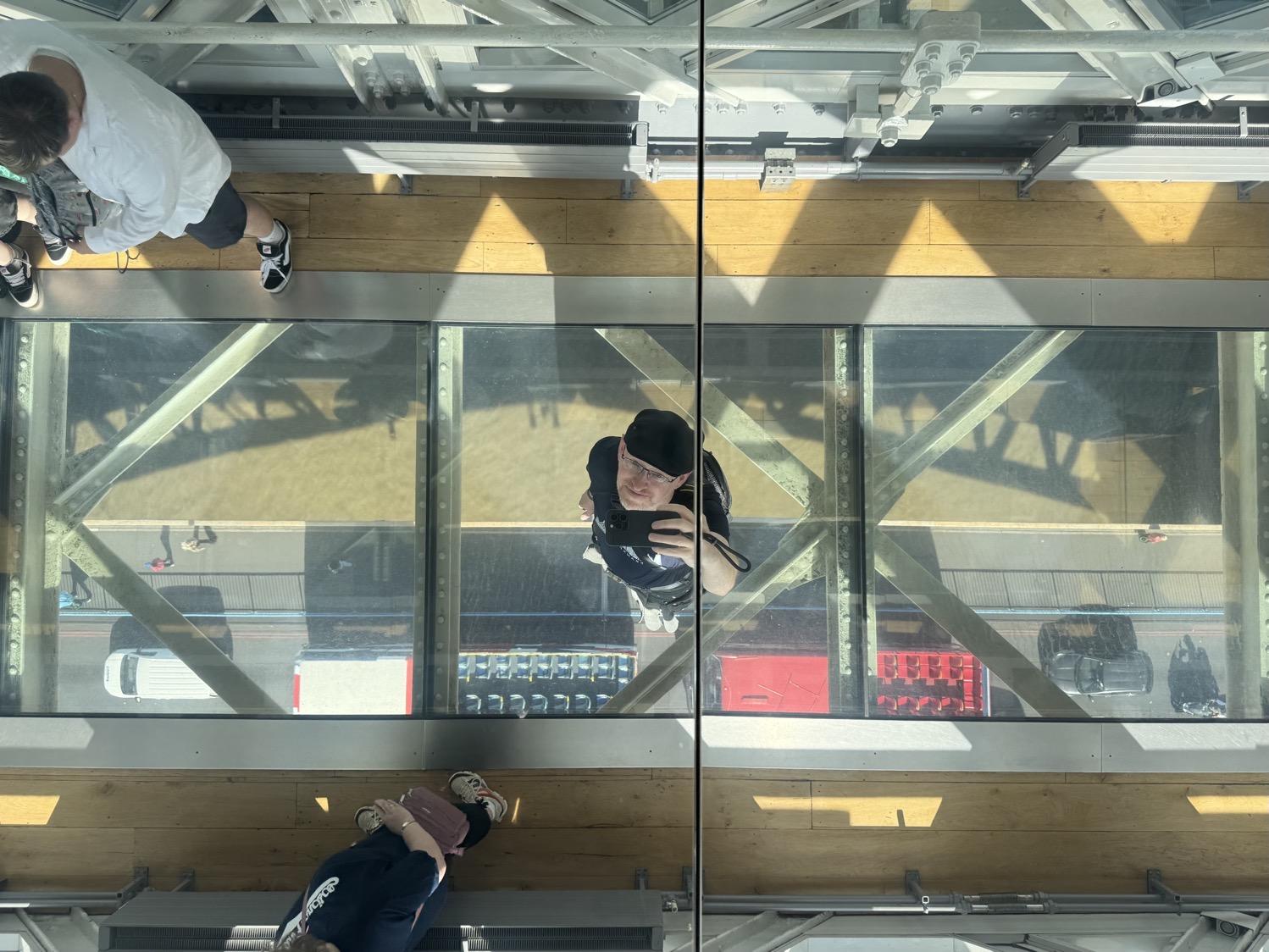 Selfie of me taken in a ceiling mounted mirror, showing me standing on a pane of glass between wooden walkways, with steel girders, a roadway, and the Thames river visible below me.
