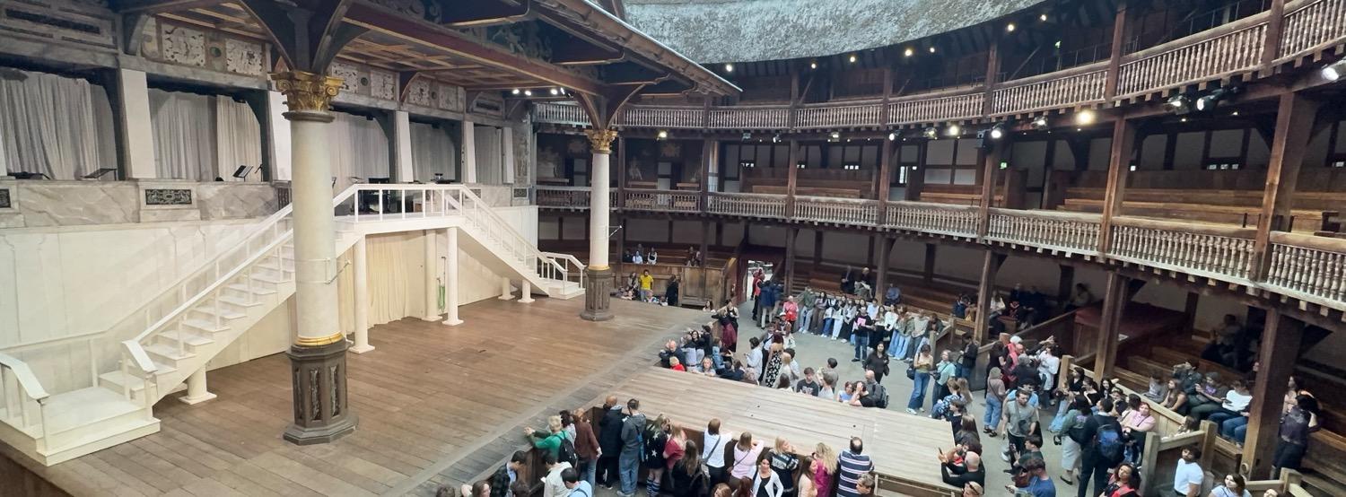 Panoramic shot of the interior of the Globe theater. The stage is to the left, with a two-level set made from white staircases and platforms. To the right of the image, three levels of curved seating areas surround a central open floor space with people standing in it.