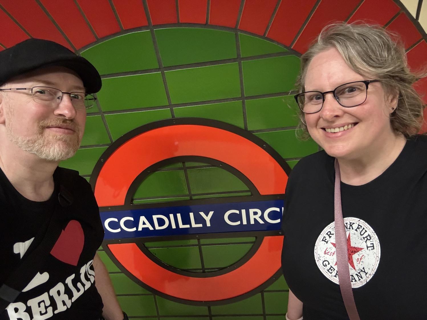 Selfie of my wife and I standing in front of a tile wall with the London Underground circle logo and the 'Piccadilly Circus' station name.