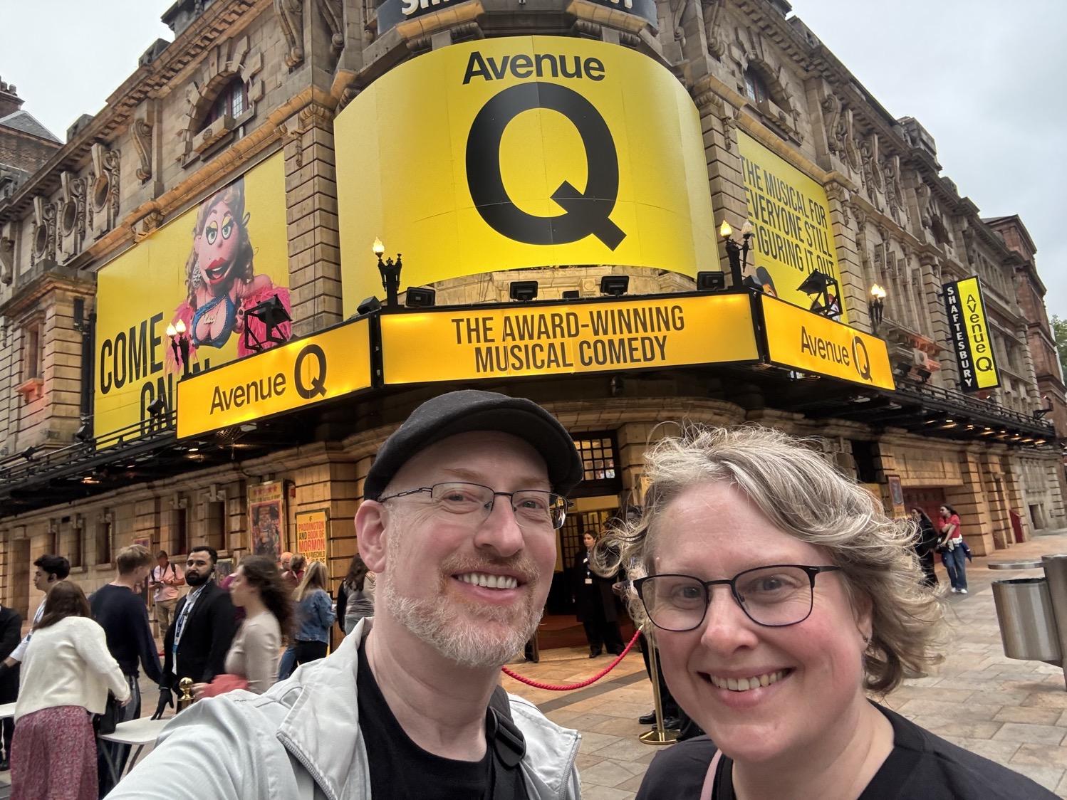 Selfie of my wife and I in front of a fancy stone building with large yellow marquee signs advertising Avenue Q.