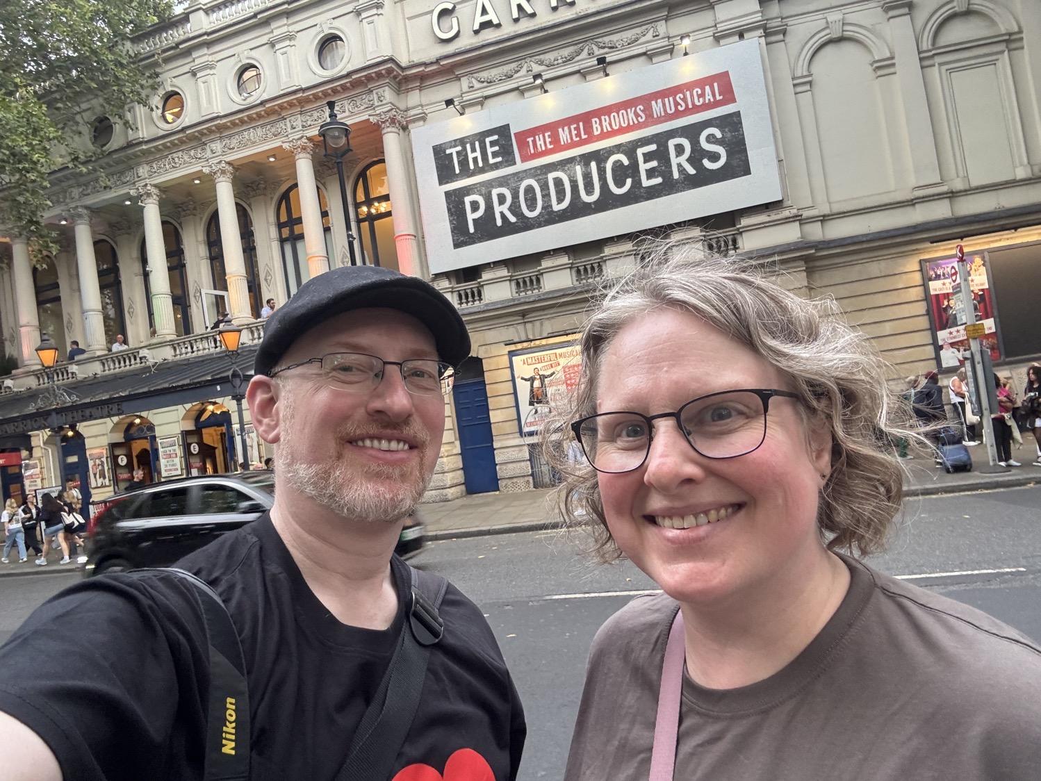 Selfie of my wife and I in front of a theater with a large black-and-red billboard advertising the Mel Brooks musical The Producers.