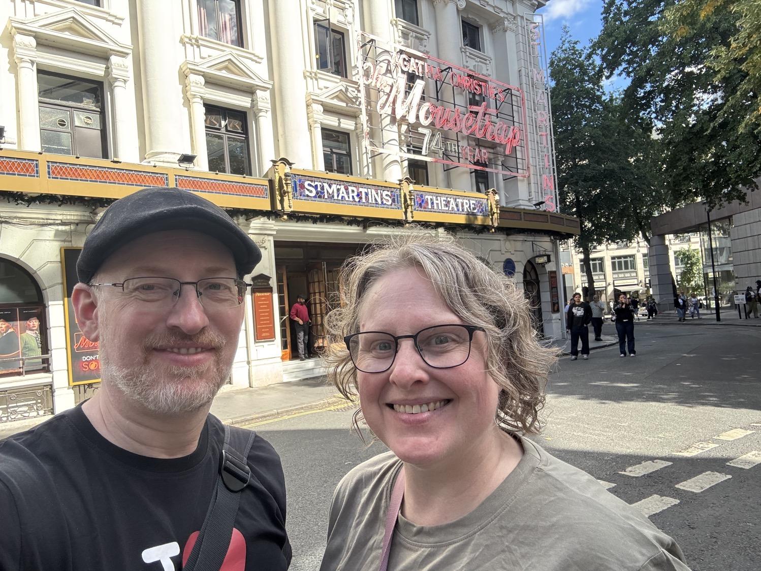 Selfie of my wife and I in front of the St. Martins theater with its neon marquee advertising The Mousetrap in it's 74th year.