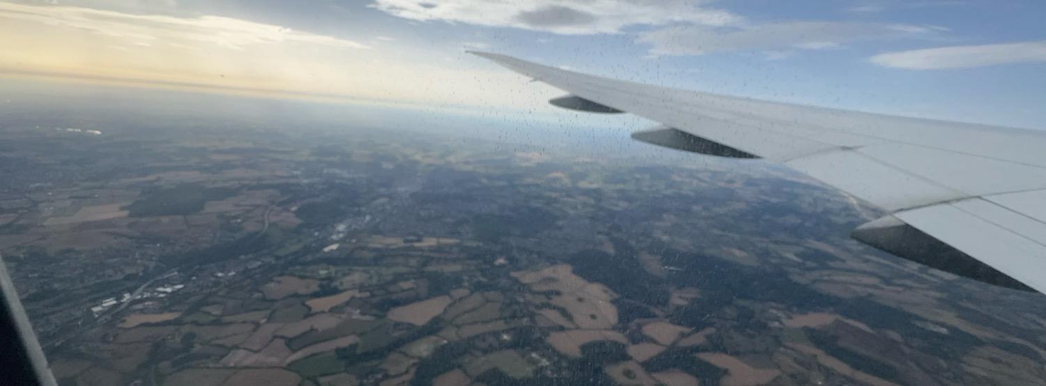 Panoramic shot of the English countryside, broken up in an irregular patchwork separated by hedges, as seen from an airplane window with the airplane wing visible to the right side of the image.
