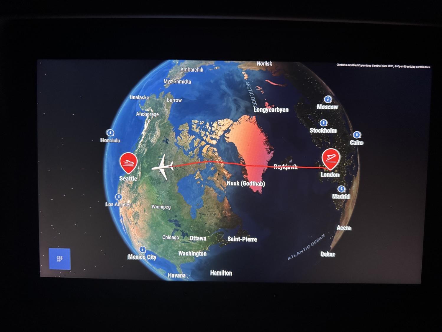 An airplane seatback screen showing a globe with an airplane flying along a route line from London over Greenland and Canada and ending at Seattle.