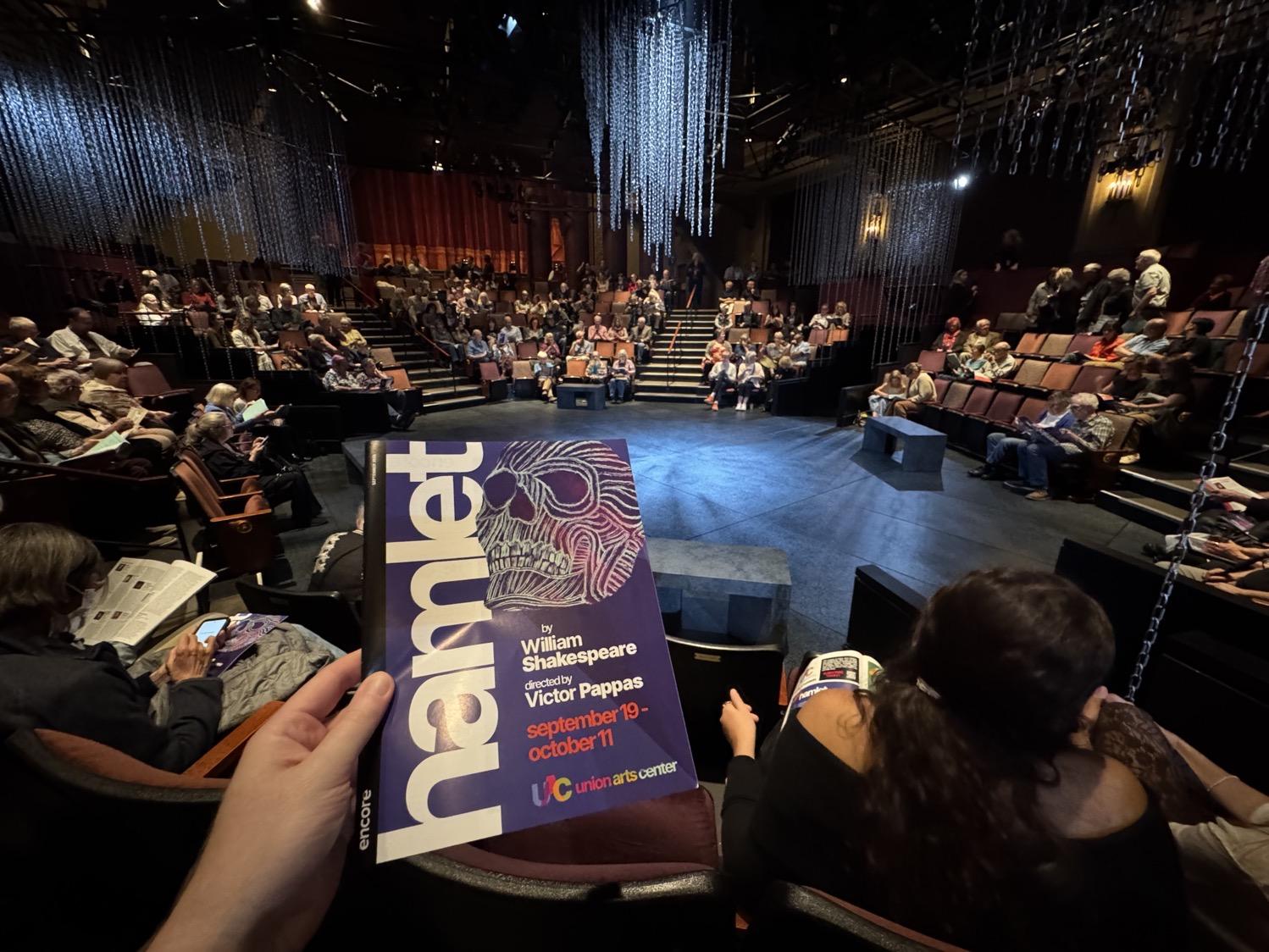 My hand holding the program for Hamlet, in a small theater with seats all the way around the stage, small benches on the stage, and chains hanging from the ceiling.