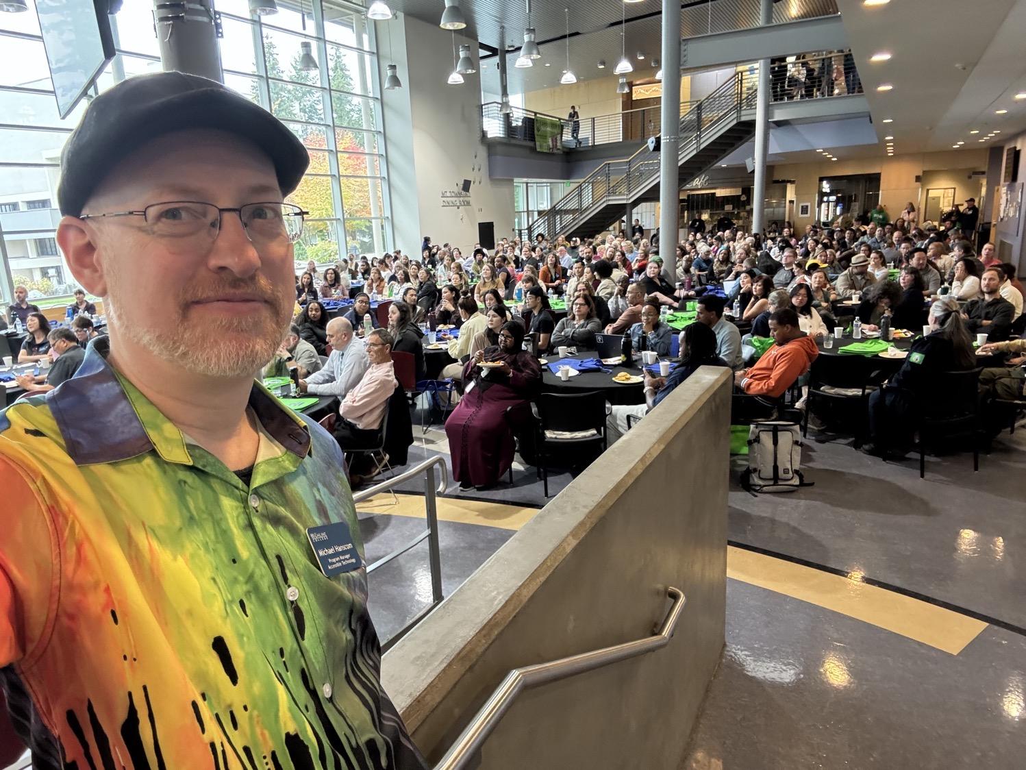 Selfie of me wearing a rainbow button-up shirt in front of a large room filled with people sitting at round tables.