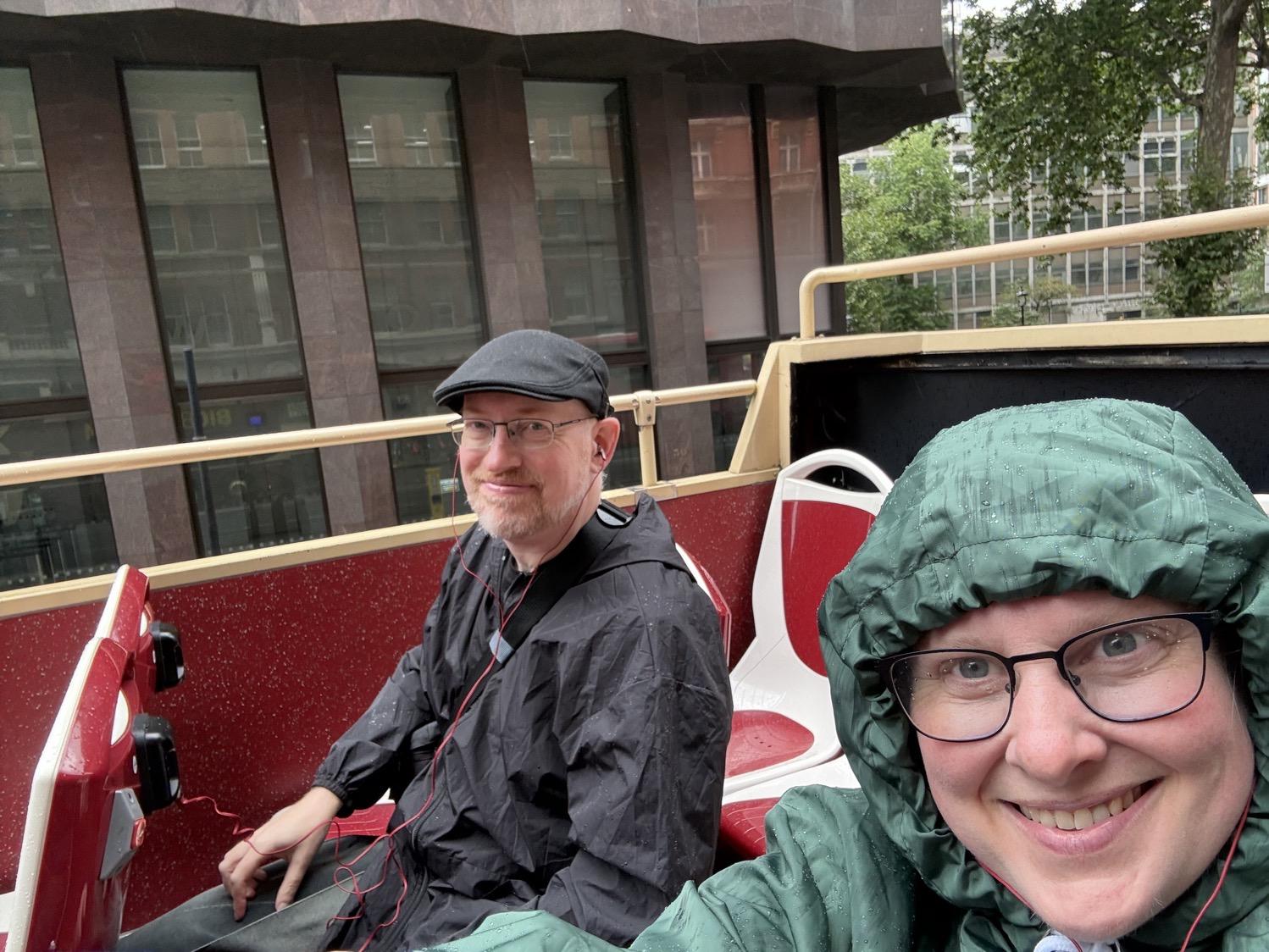 Selfie of my wife and I, both wearing obviously rain-streaked raincoats, sitting in chairs in the open section of a damp tour bus.