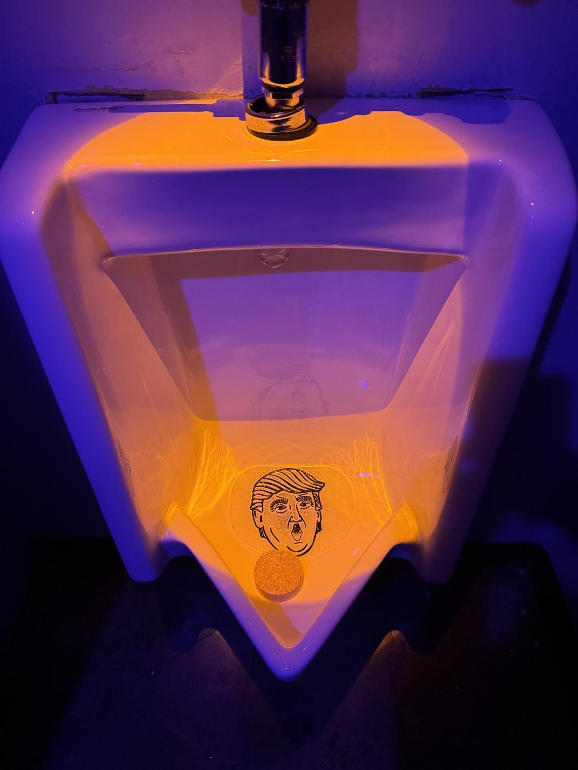 IMG_7297.jpeg A urinal lit in purple and orange, with a splash guard printed with Trump's face.