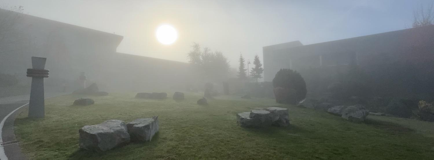 IMG_7301.jpeg In a panoramic shot, a grassy lawn with randomly placed angular boulders and a stone pillar on the left side is seen with trees and angular buildings a short distance away, all shrouded in heavy fog, with the sun trying to shine through near the upper center of the shot just above one of the buildings. It’s very mysterious and otherworldly.